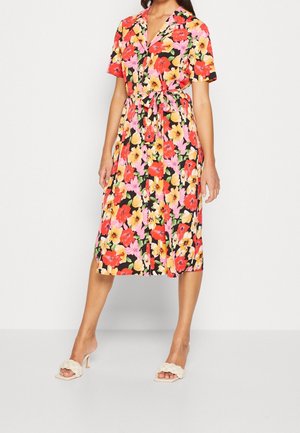 Vrouw draagt een bloemenjurk tot op de knie met korte mouwen, bedrukt met rode, gele en roze bloemen, gestrikt in de taille, gecombineerd met witte sandalen met hak.