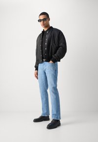 Veste bomber noire, boutons-pression argentés, polo noir, jeans bleu clair et mocassins noirs avec des accents dorés, sur un fond neutre.