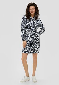 Navy- und weiß gemustertes Hemdkleid mit langen Ärmeln, Knopfleiste, Kragen und einem Bindegürtel, kombiniert mit weißen Sneakers.