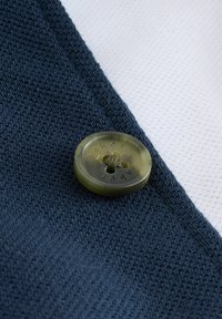 Jūras spēku adījuma audums ar gludu tekstūru, kuram ir apaļa zaļa poga ar strukturētu virsmu un iegravētu rakstu. Baltā fona.