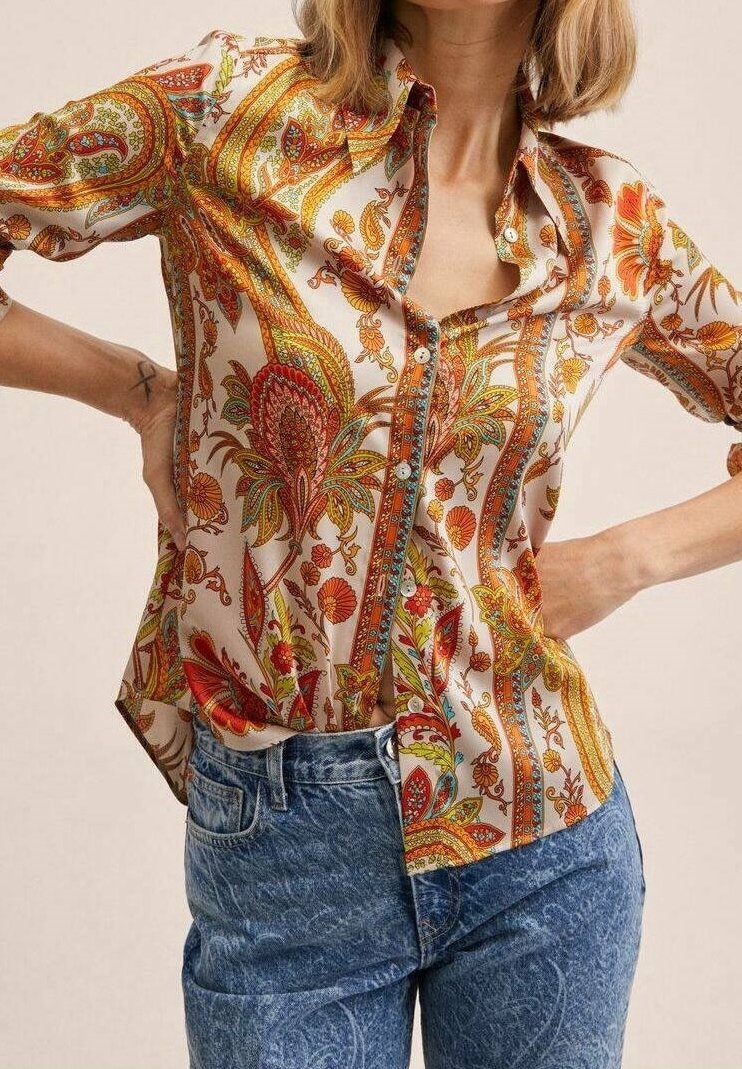 Femme portant une chemise à manches longues boutonnée avec des motifs paisley orange et un jean taille haute bleu, les mains posées sur les hanches.