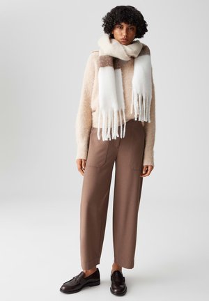 Beiger, flauschiger Pullover, braune weite Hosen und ein weiß-braun gestreifter Schal mit Fransen. Braune Loafers runden das Outfit ab.