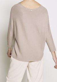 Lichtbeige trui met een losse pasvorm, driekwartmouwen, geribbelde textuur en een ronde achterkant met een kleine knoopdetail.