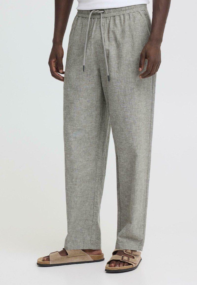 Άνδρας που φοράει φαρδιά, ανοιχτό γκρι λινά παντελόνια με κορδόνι και ταμπά σουέντ σανδάλια με διπλά λουριά, στέκεται μπροστά σε απλό φόντο.