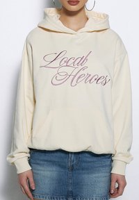 Kremowa sweatshirt z kapturem z bordowym haftem "Local Heroes", kangurzą kieszenią i ściągaczami na rękawach, zestawiona z dżinsową spódnicą.