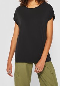 Černé tričko s krátkým rukávem a kulatým výstřihem, vyrobené z měkké látky a s volným střihem. Nosené s volnými olivovými cargo kalhotami a vrstvenými prsteny.