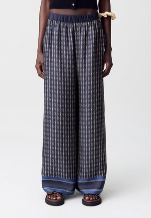 Pantaloni a gamba larga blu navy con un motivo a catena dorata, abbinati a sandali scuri, indossati da una persona in piedi davanti a uno sfondo neutro.