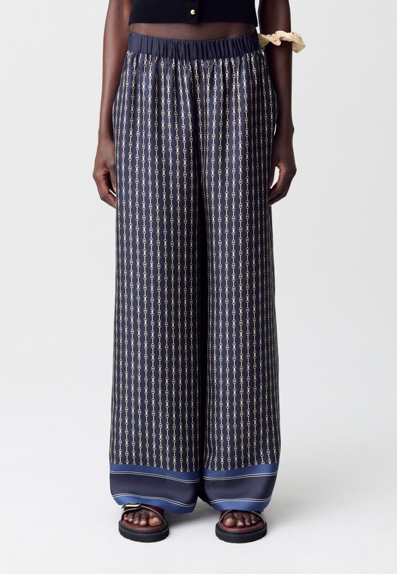 Pantalones de pierna ancha en color marino con un patrón de cadenas doradas, combinados con sandalias oscuras, usados por una persona de pie sobre un fondo neutro.