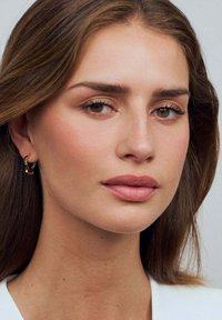 En närbild av en person med långt brunt hår, neutral sminkning och guld-hängande örhängen, mot en ljus bakgrund.