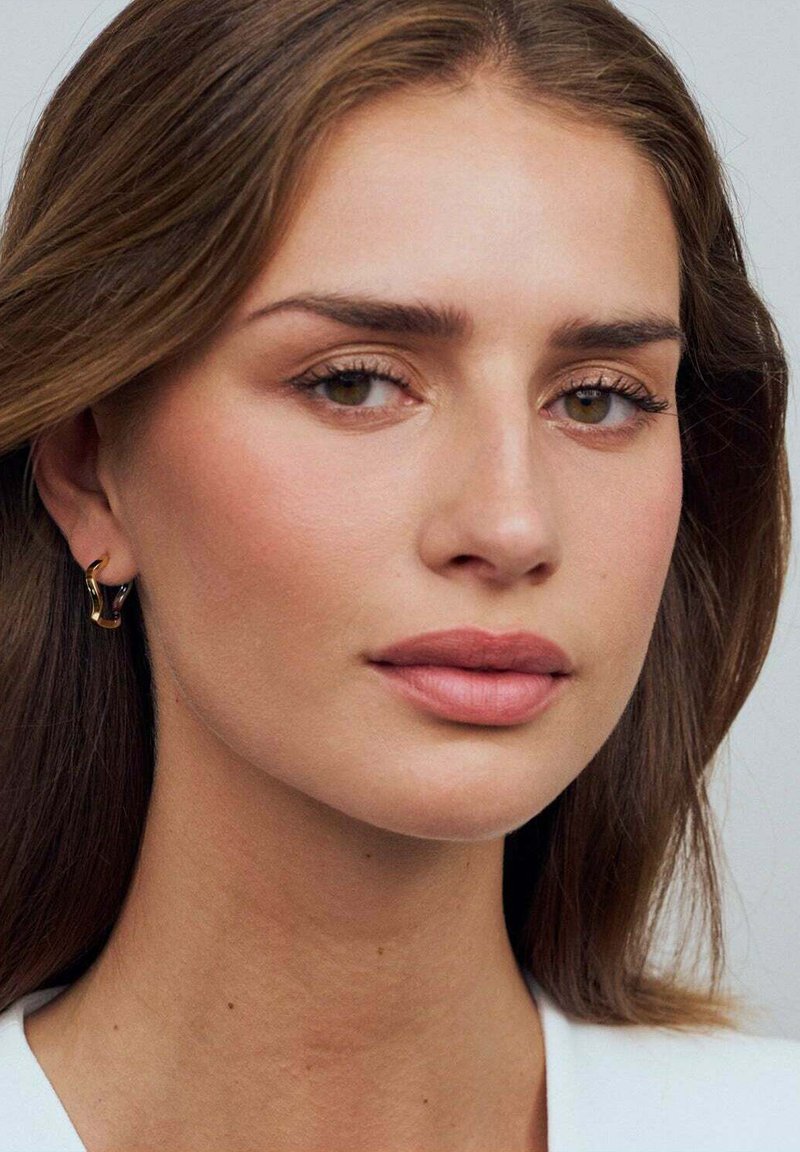 En närbild av en person med långt brunt hår, neutral sminkning och guld-hängande örhängen, mot en ljus bakgrund.