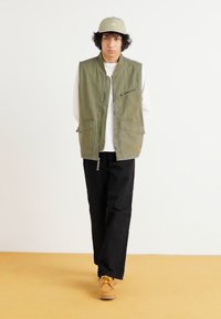 Olive green sleeveless vest with two front pockets and side zipper details; paired with a white long-sleeve shirt and black pants.
