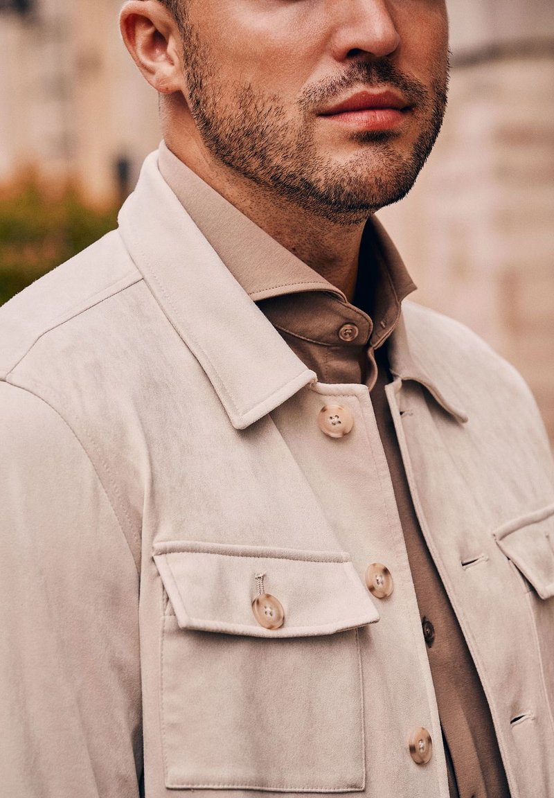 Lichtbeige suède jack met een kraag, grote borstzakken en knoopsluitingen, gecombineerd met een tankleurig hemd met knopen eronder.