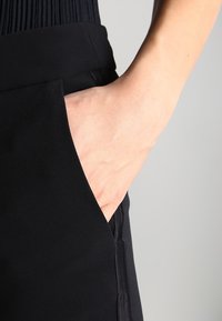 Svarta skräddarsydda byxor med en slät textur, med en sidoficka och en hand som vilar i den, vilket framhäver plaggets eleganta design.