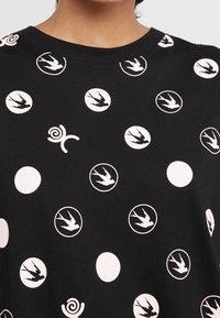 Czarny T-shirt wykonany z bawełny, z okrągłymi i spiralnymi białymi wzorami, w tym sylwetkami ptaków oraz solidnymi okręgami.