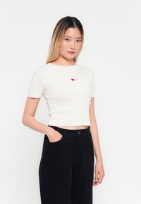 Korte mouwen, cropped, geribde t-shirt in off-white met rode hartborduring op de borst, gecombineerd met hooggewaardeerde donkere broeken.