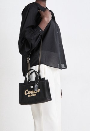 Person iført en sort skjorte med knapper og hvide bukser, der holder en sort designerhåndtaske med "Coach New York" broderet i beige.