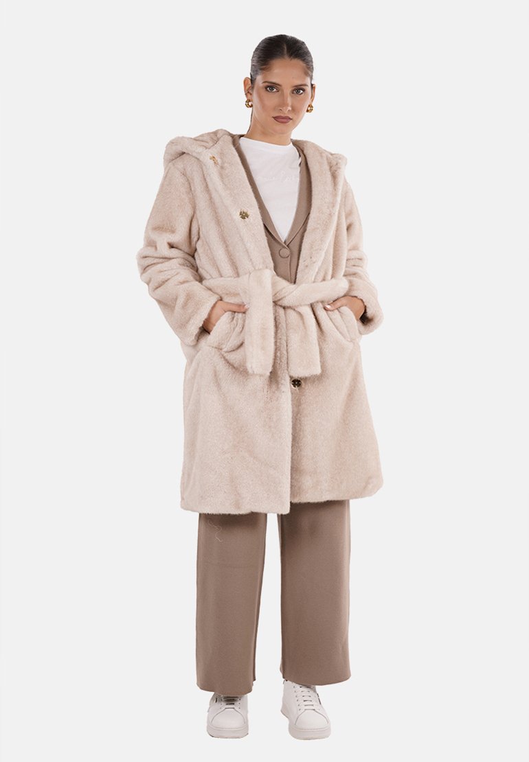 Cappotto in pelliccia ecologica color beige chiaro, con cappuccio, cintura in vita e grandi tasche. Indossato su un top crema e pantaloni marroni con sneakers.