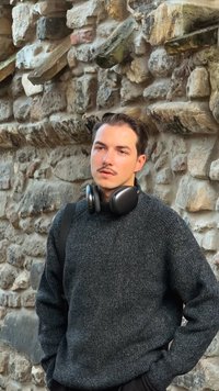 Person wearing a dark gray knit sweater with a textured finish, black over-ear headphones, set against a stone wall background.