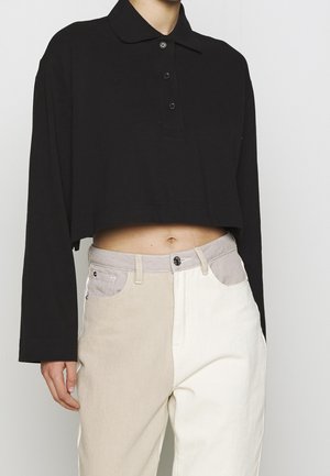 Person trägt ein schwarzes, langärmliges, kurzes Poloshirt kombiniert mit hochtaillierter zweifarbiger Hose in Beige und Weiß.