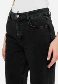 Ingrandimento di una persona che indossa jeans neri a vita alta e una canotta bianca a coste, con la mano destra poggiata di lato.