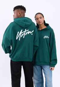 Prevelike zelene kapuce z belim logotipom na hrbtu. Ima kenguru žep, kapuco z vrvico in rebraste manšete. Unisex oblikovanje.