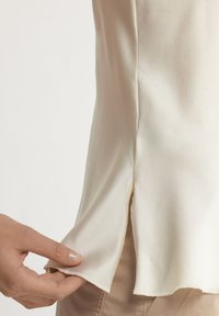 Sidenärm-lös topp i off-white, med en sidoflik och en mjuk, slät textur. Längden på fållen verkar något rundad med fina sömnadsdetaljer.