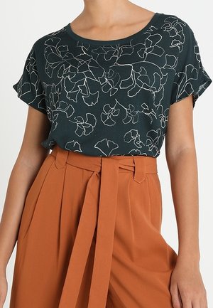 Kobieta ubrana w czarną bluzkę z krótkim rękawem z białymi kwiatowymi konturami, włożoną w wysokowasne, rdzawe pomarańczowe spodnie z materiałowym paskiem.