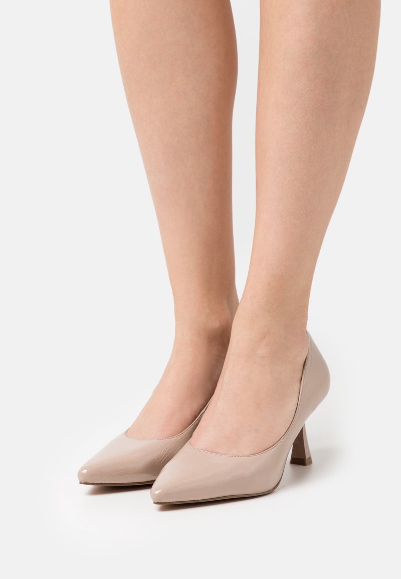 Femme portant des chaussures en cuir verni couleur nude à bout pointu et talon moyen, debout sur une surface blanche unie.