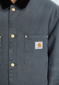 Graue Arbeitsjacke aus strapazierfähigem Stoff, mit einem Stehkragen aus Cord, zwei frontalen Taschen mit goldener Hardware und einem Carhartt-Logo-Patch.