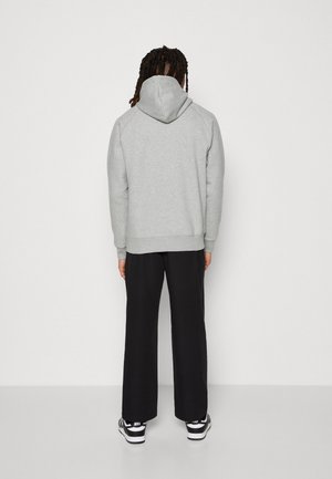 Person mit geflochtenem Haar, die einen hellgrauen Hoodie, schwarze Hosen mit weitem Bein und schwarz-weiße Nike-Sneaker trägt und einer schlichten Wand zugewandt steht.
