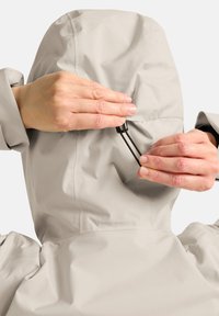 Veste imperméable gris clair avec capuche. Des mains ajustent le cordon de la capuche. Le matériau semble lisse et légèrement brillant.