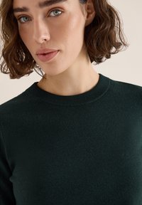 Dunkelgrüner Pullover mit rundem Halsausschnitt, geripptem Kragen und glatter Textur. Der Stoff wirkt weich und leicht.