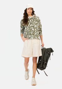 Olivenfarvet sweatshirt med camouflagemønster, beige shorts, lyse sneakers og en mørkegrøn rygsæk med lynlåse og stropper.