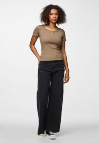 Mlada žena s kovrčavom kosom, odjevena u smeđu kratku majicu, crne hlače širokih nogavica i crno-bele tenisice, stoji na bijeloj pozadini.