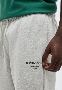 Κοντινό πλάνο ενός ατόμου που φοράει ανοιχτό γκρι παντελόνι φόρμας Björn Borg με κορδόνι στη μέση και μαύρο λογότυπο στο μηρό, με το χέρι χαλαρό στο πλάι.