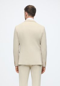 Šviesiai rudos spalvos pritaikyta švarkas su lygiu paviršiumi, siauru kirpimu, vienu įpjovimu nugaroje ir auksinės spalvos sagutėmis ant rankovių. Tik atbulinis vaizdas.