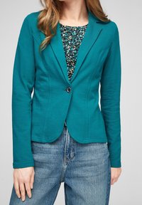 Blazer stretch sarcelle avec un seul bouton noir, coupe ajustée, ourlet incurvé et manches longues, superposé à un haut à motif floral.