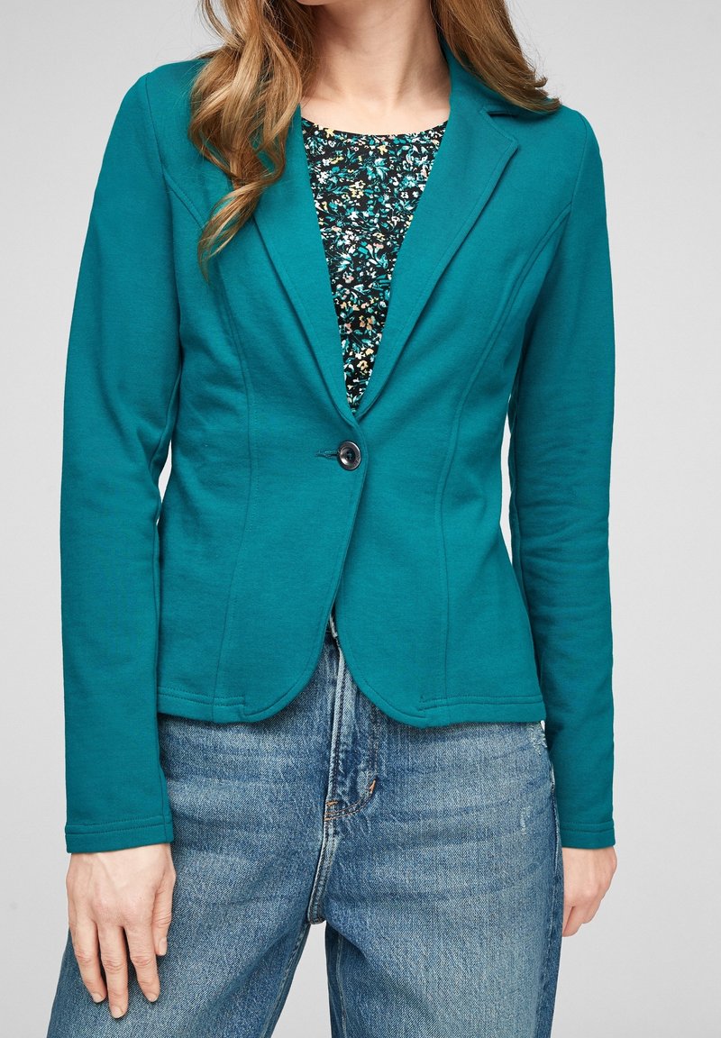 Blazer stretch sarcelle avec un seul bouton noir, coupe ajustée, ourlet incurvé et manches longues, superposé à un haut à motif floral.