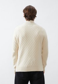 Man med mellanhårig brunt hår bär en krämfärgad ribbad tröja och svarta byxor, står vänd mot en vit vägg.