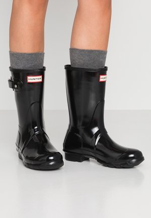 Personne portant des bottes de pluie Hunter noires brillantes mi-mollet avec des chaussettes grises, debout sur un sol blanc devant un fond uni.