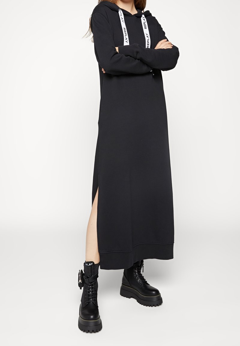 Femme portant une longue robe à capuche noire avec une fente sur le côté et des bottes noires épaisses à lacets avec une étiquette de marque.