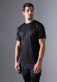 Črna kratkotrajna športna majica s kratkimi rokavi, gladka tekstura, okrogla vrata, majhna oznaka na levem prsnem delu, v kombinaciji s črnimi hlačami. Preprost dizajn.