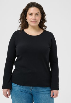 Femme aux cheveux bruns bouclés portant un haut à manches longues noir et un jean bleu, debout devant un fond clair uni.