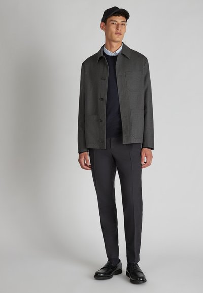 Grau wollene Jacke mit Knopfverschluss und zwei Fronttaschen, getragen über einem dunklen Pullover und kombiniert mit schmalen schwarzen Hosen und Schuhen.