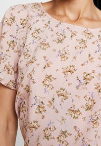 Blus med blommönster i mjukt rosa tyg med små bruna, gröna och lila blommor. Kort ärm och rund halsringning med slät textur.