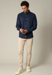 Marineblauw overhemd met lange mouwen, gecombineerd met lichtbeige broek en crèmekleurige sneakers. Het overhemd heeft een borstzak en een klassieke kraag.