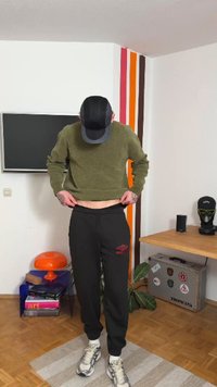 Alyvuogių žalios spalvos trumpas megztinis, juodos joggerinės kelnės su raudonu logotipu, šviesūs sportbačiai, stovintis kambaryje su medinėmis grindimis ir spalvingomis sienų juostomis.