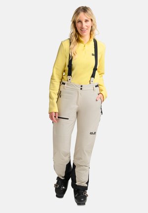Beige Skihosen mit schwarzen Akzenten und Trägern, ausgestattet mit Reißverschlusstaschen und einem Logo. Kombiniert mit einem gelben Fleece-Oberteil.