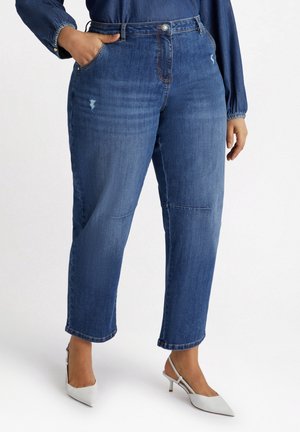 Vrouw draagt blauwe spijkerbroek met lichte slijtage, gecombineerd met witte slingback-hakken met puntige neus, op een witte achtergrond.