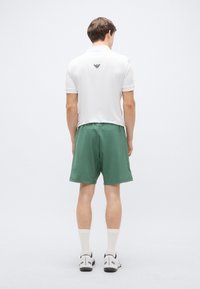 Hvit polotrøye med en svart logo på baksiden, kombinert med grønne shorts. Modellen har på seg hvite sokker og grå treningssko.